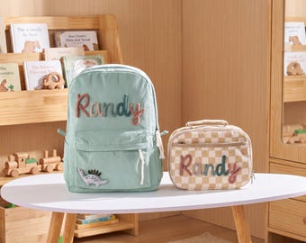 Könnte beinhalten: Ein hellgrüner Cord-Rucksack und eine beige karierte Lunchtasche, beide mit dem Namen "Randy" in farbenfrohen, strukturierten Buchstaben personalisiert. Der Rucksack hat einen Dinosaurier-Aufnäher. Die Artikel stehen auf einem weißen Tisch.