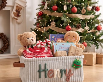 May include: A light gray corduroy storage basket with the name Henry embroidered in green, cream, and brown yarn. The basket features brown faux leather handles and a small green Christmas tree patch, filled with a teddy bear, books, and gifts.