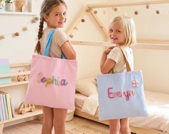 Könnte beinhalten: Zwei rechteckige Tragetaschen aus Seersucker-Stoff. Eine Tasche ist hellrosa mit dem Namen Sophia in bunten, erhabenen Buchstaben. Die andere Tasche ist hellblau mit dem Namen Evelyn in rosa erhabenen Buchstaben und einer kleinen Blumenapplikation.