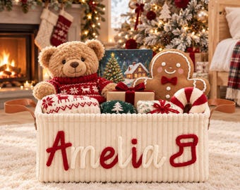 May include: A cream-colored, ribbed fabric storage basket with brown leather handles, featuring the name Amelia and a stocking icon in red and white felt lettering. The basket is filled with a teddy bear, a gingerbread man, and gifts.