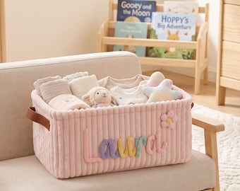 May include: A rectangular, light pink corduroy storage basket featuring the name Laura in multi-colored yarn lettering and a small flower appliqué. The basket is filled with folded baby clothes, a plush lamb toy, and a star-shaped pillow.