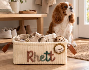 May include: A rectangular cream-colored corduroy storage basket featuring the name Rhett embroidered in brown and green yarn. The basket includes brown faux leather side handles and a plush lion face appliqué on the front right corner.