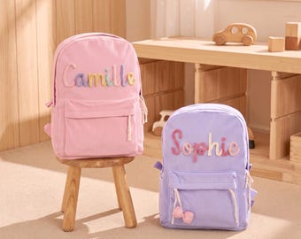 Könnte beinhalten: Ein rosafarbener Kordrucksack mit dem Namen Camille in bunten Buchstaben und ein violetter Kordrucksack mit dem Namen Sophie in bunten Buchstaben, jeweils mit einer Vordertasche, Reißverschlüssen und einem kleinen karierten Schleifendetail.