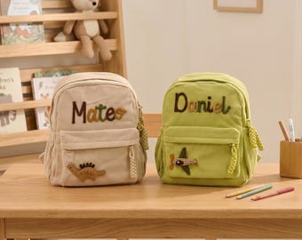 Könnte beinhalten: Zwei Cord-Rucksäcke, einer beige und einer grün, stehen auf einem Holztisch. Der beige Rucksack hat den Namen "Mateo" aufgestickt und ein Dinosaurier-Applikation. Der grüne Rucksack hat den Namen "Daniel" und eine Flugzeug-Applikation. Beide haben Reißverschlusstaschen.