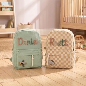 May include: Two corduroy backpacks. The left one is sage green with the name Daniel embroidered in brown and green, featuring a small airplane patch. The right one has a beige and white checkerboard pattern with the name Rhett and a dinosaur patch.