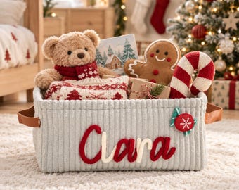 May include: A light gray corduroy storage basket with brown faux leather handles, featuring the name Clara in red and white script. The basket is filled with a teddy bear, a gingerbread man, a candy cane, and a gift box.