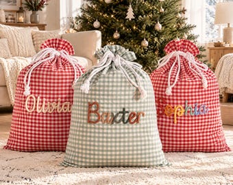 May include: Three fabric drawstring gift sacks with a gingham pattern. Two sacks are red and white, and one center sack is sage green and white. Each sack features a personalized name in raised lettering: Olivia, Baxter, and Sophia.