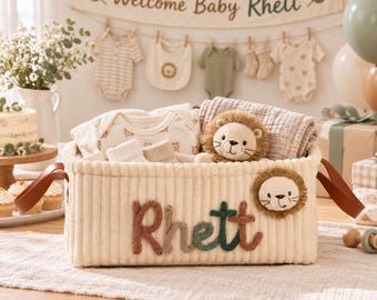 May include: A cream-colored corduroy storage basket featuring the name Rhett in multi-colored yarn lettering and a plush lion face patch. The basket contains baby clothing, a small plush lion toy, and a folded muslin blanket.
