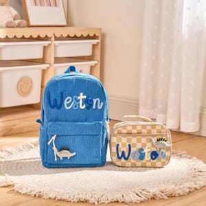 May include: A blue corduroy backpack and matching lunch bag. The backpack features the name "Weston" in blue and white letters, with a dinosaur applique. The lunch bag has a checkered pattern and the name "Weston".