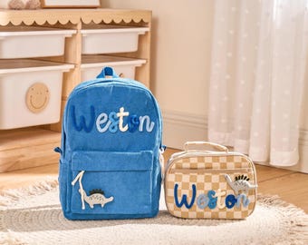 Könnte beinhalten: Ein blauer Cord-Rucksack und eine passende Lunchtasche. Der Rucksack trägt den Namen "Weston" in blauen und weißen Buchstaben mit einem Dinosaurier-Applikation. Die Lunchtasche hat ein Karomuster und den Namen "Weston".