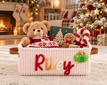 May include: A light pink corduroy storage basket with brown faux leather handles. The name Riley is embroidered in red, orange, yellow, and white yarn on the front. A green felt wreath patch is attached to the side of the basket.