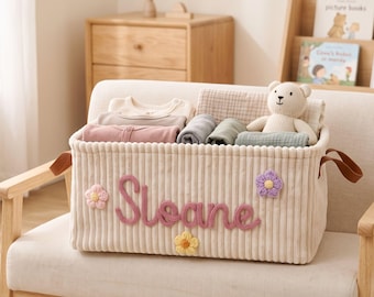 May include: A cream-colored corduroy storage basket featuring the name Sloane in pink yarn lettering, accented with three small crocheted flowers in pink, yellow, and purple. The basket contains folded baby clothing and a small knit teddy bear.
