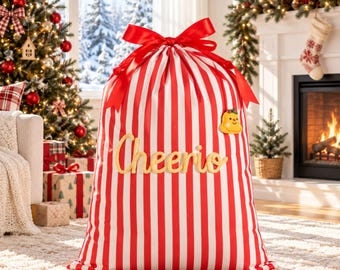 May include: A large drawstring gift sack featuring vertical red and white stripes, tied at the top with a red satin ribbon. The word Cheerio is written in gold script across the front, accompanied by a small yellow bell-shaped ornament.