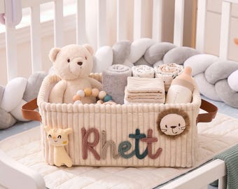 May include: A rectangular beige corduroy storage basket featuring the name Rhett in raised, multi-colored knit lettering. The basket is decorated with a plush giraffe and a lion face, and contains a teddy bear, a baby bottle, and folded cloths.