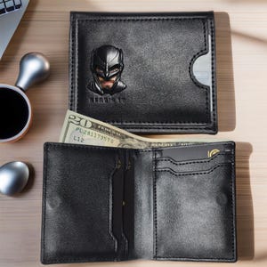 May include: A black leather wallet with a Batman logo on the front. The wallet is open to reveal multiple card slots and a money clip.
