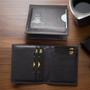 May include: A black leather wallet with a Batman logo and the text "Hero's ID". The wallet is open and has three credit cards inserted.