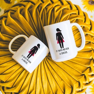 May include: Two white ceramic mugs with the text "IT WAS NEVER A DRESS" and a graphic of a person in a dress. The mugs are on a yellow, textured cushion with a sunflower print background.