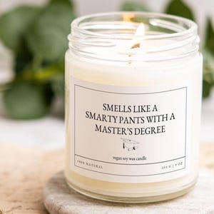 Puede incluir: Una vela en frasco de vidrio transparente con cera blanca y una mecha encendida. La etiqueta presenta el texto "SMELLS LIKE A SMARTY PANTS WITH A MASTER'S DEGREE" sobre un icono de birrete, "vegan soy wax candle", "100% NATURAL", "255 G".