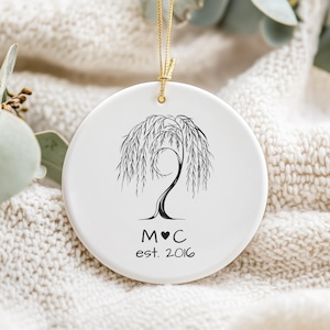 May include: A white ceramic ornament with a black weeping willow tree design. The ornament has a gold string for hanging and features the initials "M♥C" and the year "est. 2016" in black text. The background is a soft, textured white fabric.