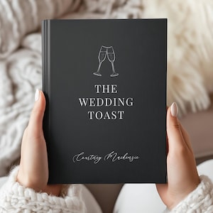 May include: A dark gray book titled "The Wedding Toast" with two champagne glasses illustrated above the title. The author's name, Courtney Mackenzie, is written in cursive below the title. The book is held by a person wearing a white sweater.