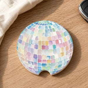 May include: A circular car coaster featuring a multi-colored mosaic pattern of small squares in shades of blue, pink, yellow, and purple. The coaster has a smooth, flat surface and a small semi-circular notch cut out from the bottom edge for easy removal.