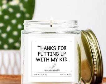 May include: A clear glass jar candle with a gold lid, featuring a white label with the text "THANKS FOR PUTTING UP WITH MY KID." The candle is 4 oz and made of soy wax. The candle is on a wooden surface.