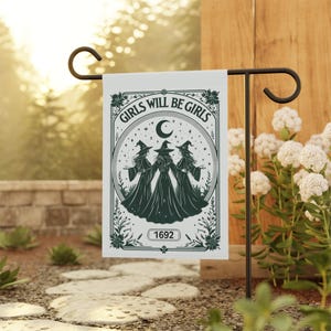May include: A white garden flag with a black wrought iron stand. The flag features a circular design with three witch figures, a crescent moon, and the text "GIRLS WILL BE GIRLS" and "1692". The flag is displayed outdoors.
