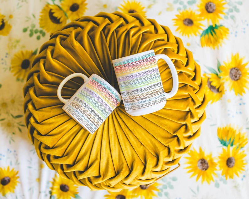 May include: Two white coffee mugs with rainbow-colored embroidered patterns rest on a yellow, pleated cushion. The mugs have a simple, curved handle. The background is a floral pattern with sunflowers.
