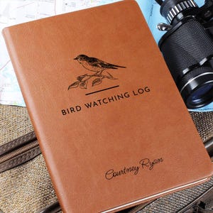 May include: A brown leather-bound "Bird Watching Log" notebook. The cover features a detailed illustration of a bird perched on a branch, with the text "Bird Watching Log" below. The name "Courtney Ryan" is printed at the bottom.