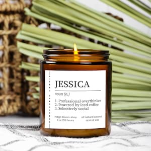 Puede incluir: Una vela encendida en un frasco de vidrio marrón con una etiqueta blanca que dice "JESSICA" y una definición. La vela es de 255g y está hecha con cera de albaricoque de coco natural. El fondo incluye hojas verdes y una cesta tejida.