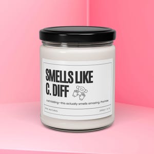 Puede incluir: Un tarro de vela blanco con tapa negra y una etiqueta blanca que dice "SMELLS LIKE C. DIFF" con un gráfico de una mascarilla facial y dos manos enguantadas. La etiqueta también dice "Just kidding-this actually smells amazing. Promise" y "100% NATURAL".