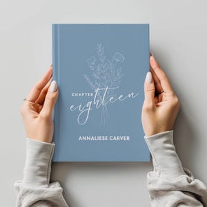 May include: A blue journal with the text "Chapter Eighteen" and the name "Annaliese Carver" in white script. A floral illustration is above the text. The journal is held by two hands.