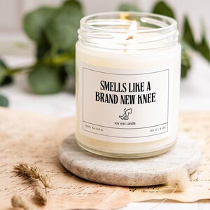 Könnte beinhalten: Eine weiße Sojawachskerze in einem Glasgefäß mit einem schwarzen Etikett, auf dem "Smells Like A Brand New Knee" steht. Das Etikett enthält auch eine kleine Illustration eines Beins mit einem Verband am Knie. Die Kerze ist 255 g schwer und 100% natürlich.