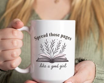 Könnte beinhalten: Weißer Keramikbecher mit dem Text "Spread those pages like a good girl" und einer Illustration eines offenen Buches mit Blättern. Der Becher wird von einer Person gehalten, die einen grünen Pullover trägt.