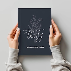 Puede incluir: Un diario azul oscuro con un diseño floral blanco y el texto "Chapter Thirty" en una fuente cursiva. El nombre "Annaliese Carver" está impreso debajo del diseño floral.