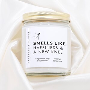 Könnte beinhalten: Ein Glas-Kerzenglas mit goldenem Deckel. Auf dem Etikett steht "SMELLS LIKE HAPPINESS & A NEW KNEE". Die Kerze ist handgegossen und aus Kokos-Aprikosenwachs hergestellt. Sie wiegt 255 Gramm und hat eine Brenndauer von 55 Stunden.