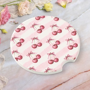 May include: A round car coaster with a white background and a pattern of pink stripes and cherries. Each cherry cluster is tied with a pink bow. The coaster has a small notch for easy removal from the car's cup holder.