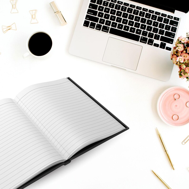 May include: A black lined notebook with a white interior is open on a white surface. A laptop computer, a cup of coffee, gold clips, and a pink dish with gold rings are also on the surface.