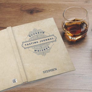 May include: A brown leather-bound journal with a blue and white decorative design. The journal is titled "Bourbon Tasting Journal Whiskey" and has the name "Stephen" printed on the cover.