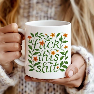 May include: White ceramic mug with the phrase "fuck this shit" in green script, surrounded by green vines and orange flowers. The mug is held by a person wearing a white sweater.