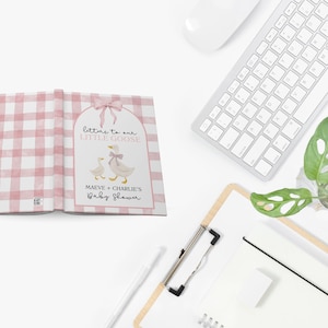 May include: A baby shower guest book with a pink gingham pattern. The cover features a watercolor illustration of two geese and the text "Letters to our Little Goose." The book is open, revealing blank pages.
