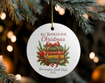 Pode incluir: Enfeite de Natal redondo branco com o texto "ALL BOOKED FOR Christmas". Apresenta uma pilha de livros com um laço vermelho, azevinho e uma pinha. O texto "Barrington Book Club 2025" também está incluído.