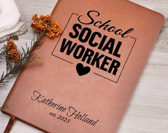 May include: A brown leather-bound journal with the words "School SOCIAL WORKER" in bold black letters, a black heart, and the name "Katherine Holland" with "est. 2025" below. A decorative sprig of orange flowers rests on the side.