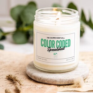 May include: A white candle in a glass jar with a label that reads "The calming scent of a colour coded spreadsheet." The label also says "Vegan Soy Wax" and "255 g | 9 oz".