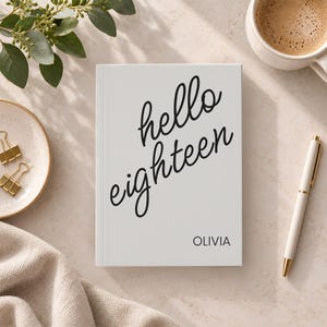 Puede incluir: Un cuaderno rectangular blanco con texto cursivo negro que dice hello eighteen y el nombre OLIVIA impreso en una fuente sans-serif simple en la parte inferior. El cuaderno tiene una cubierta lisa y está colocado verticalmente sobre una superficie.