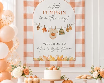 May include: A rectangular fabric backdrop featuring a white and orange buffalo plaid border. The centre displays an arched design with the text "a little PUMPKIN is on the way!" and "WELCOME TO Maeve's Baby Shower" above a row of autumn leaves.