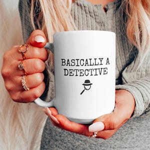May include: White ceramic mug with black text that reads "Basically a Detective" and a black silhouette of a magnifying glass with a hat on top.