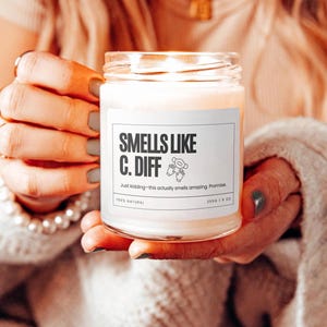 Puede incluir: Una vela blanca en un tarro de vidrio con una etiqueta blanca que dice "Smells Like C. Diff" y "Just kidding - this actually smells amazing. Promise." La etiqueta tambi&eacute;n dice "100% Natural" y "2550 | 8 oz".