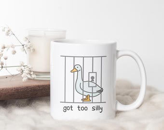 Könnte beinhalten: Weiße Keramiktasse mit einer Cartoon-Illustration einer Gans in einer Gefängniszelle. Der Text "got too silly" ist unter dem Bild gedruckt.