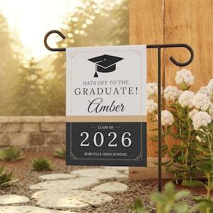 May include: A graduation garden flag with a black metal stand. The flag has a graduation cap graphic and text that reads "Hats off to the Graduate! Amber, Class of 2026, Borcelle High School."